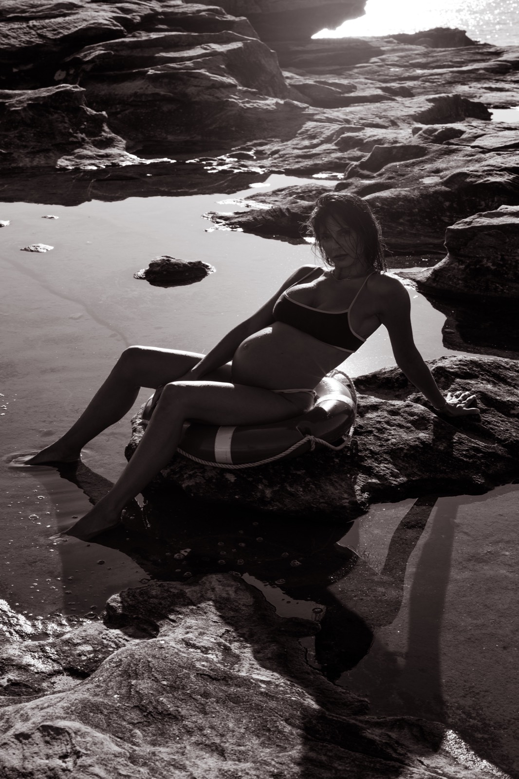 Black-and-white maternity portrait seated on a lifesaver ring beside the ocean