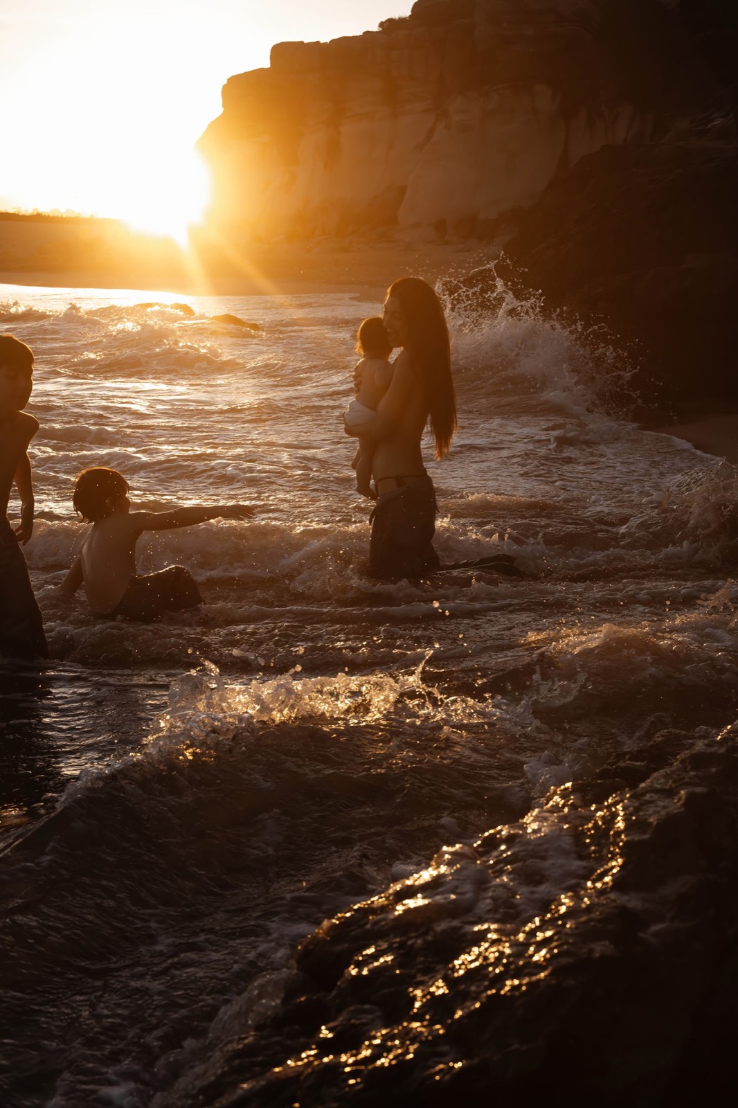 Mother holding her baby as her children play in the ocean at sunset